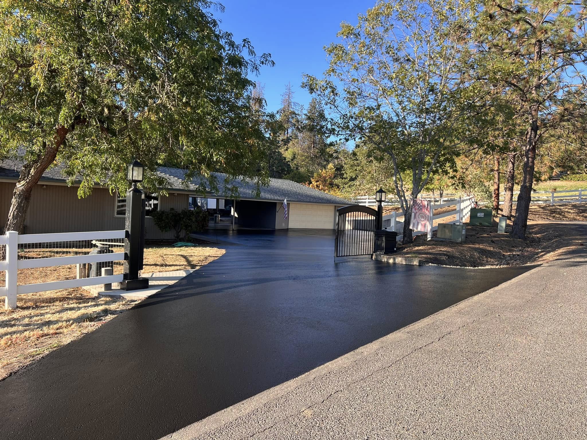 Dark asphalt driveway leading through an open gate to a home and garage, bordered by trees and a white fence.
