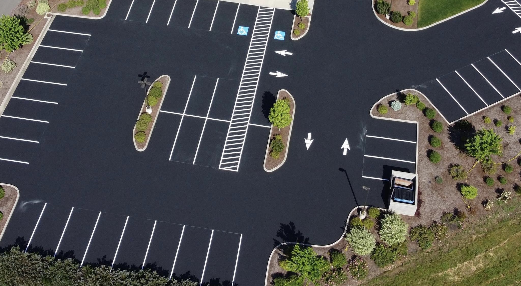 Aerial view of a freshly sealcoated commercial parking lot with bright white parking stall lines, directional arrows, ADA parking spaces, and landscaped islands throughout the property. The smooth black asphalt creates a clean, organized appearance with clearly marked traffic flow and parking areas.